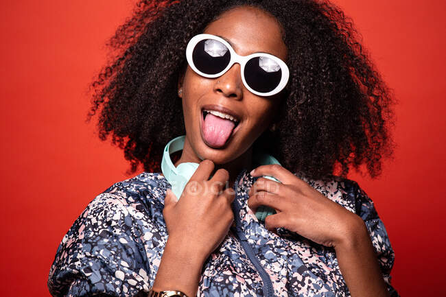 Cool joven afroamericana femenina en traje de moda y gafas de sol con auriculares inalámbricos alrededor del cuello y mostrando la lengua contra el fondo rojo - foto de stock