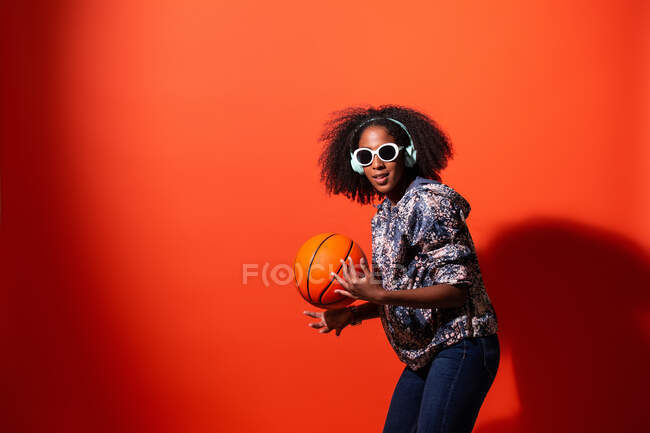 Contenido joven mujer afroamericana en traje elegante y gafas de sol escuchando música en los auriculares y jugando con la pelota de baloncesto mientras mira a la cámara contra el fondo rojo - foto de stock
