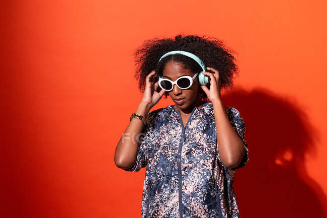 Joven mujer afroamericana alegre en traje elegante escuchando música fresca en auriculares inalámbricos y mirando hacia otro lado con gafas de sol contra la pared roja - foto de stock