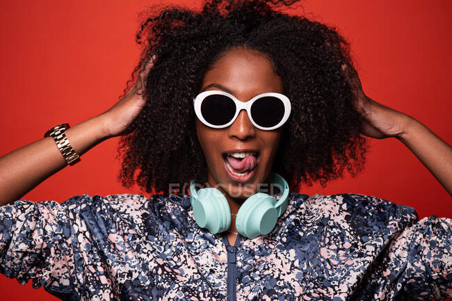 Cool young African American female in trendy outfit and sunglasses carrying wireless headphones around neck touching Afro hair and showing tongue against red background — Stock Photo