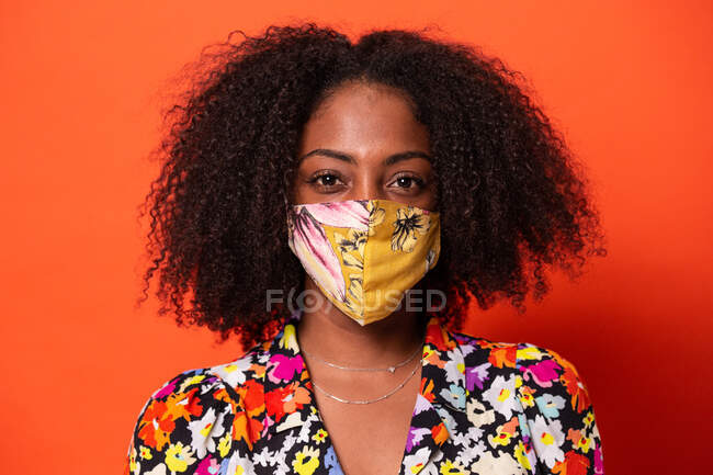 Attractive young African American female in colorful top wearing cloth face mask and looking at camera against red background — Stock Photo