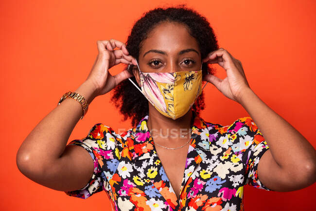 Attrayant jeune afro-américaine femelle en haut coloré mettant masque en tissu et en regardant la caméra sur fond rouge — Photo de stock