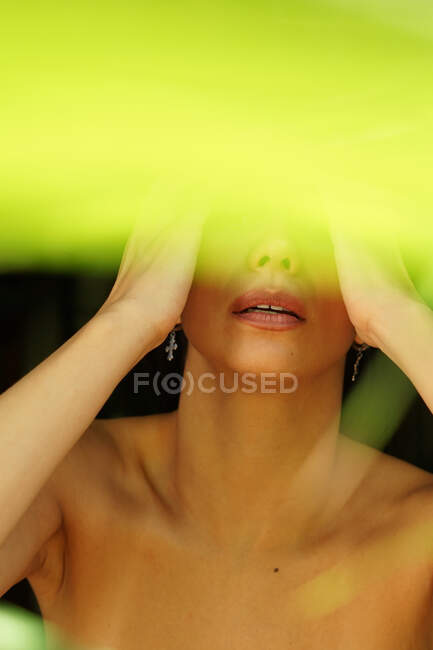 Чувствительная к растениям женщина в серьгах и макияж на губах касаясь щек за ярко-зеленым светом — стоковое фото