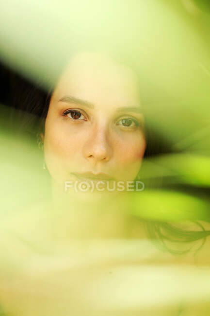 Jovem fêmea suave solitária com os olhos fechados em pé atrás de folha de planta verde colorido — Fotografia de Stock