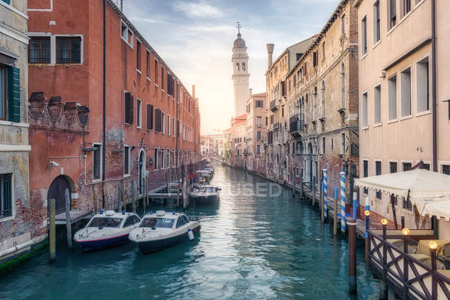 Лодки пришвартованы на водном канале между обветшалыми жилыми зданиями под закатным небом в Венеции — стоковое фото