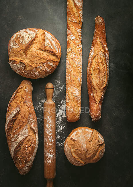 Композиція зверху з різними типізованими свіжоспеченими хлібними хлібними хлібами різних форм, розташованими біля дерев'яного штифта на чорному тлі — стокове фото