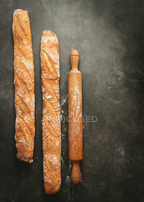 Top view composition with two artisan bread loaves placed near wooden rolling pin on black background — Stock Photo