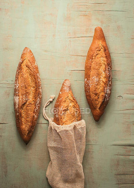 Composición de vista superior de deliciosos panes de masa fermentada artesanal crujientes envasados en bolsas de arpillera sobre fondo verde - foto de stock
