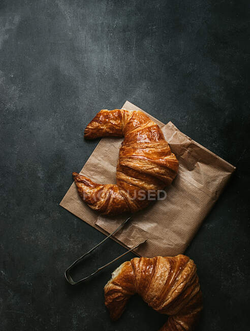 Top view composition with tasty fresh crusty croissants placed with paper bag and metal tongs on black background — Stock Photo