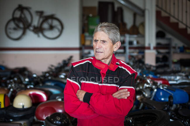 Серьезный старший механик в красной рабочей одежде, стоящий в ремонтной мастерской против поврежденных ржавых мотоциклов, отворачивающихся — стоковое фото