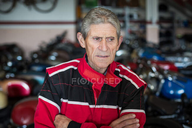 Grave anziano meccanico maschio in abito da lavoro rosso in piedi in officina di servizio di riparazione contro i motocicli arrugginiti danneggiati guardando altrove — Foto stock