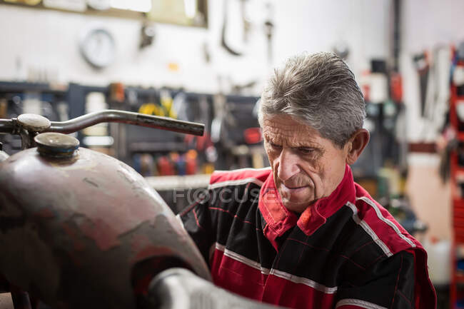 Senior männlicher Mechaniker repariert altes rostiges zerlegtes Motorrad während er in einer professionellen Werkstatt arbeitet — Stockfoto