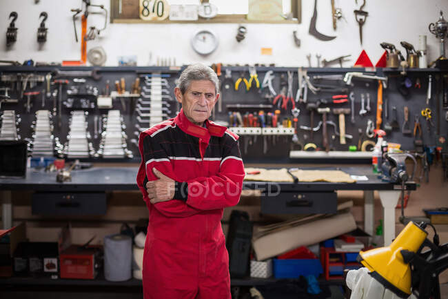 Серьезный старший механик в красной рабочей одежде, стоящий в ремонтной мастерской с профессиональными инструментами и инструментами, отводящими взгляд — стоковое фото
