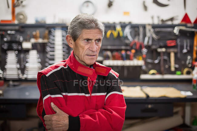 Ernsthafter Senior-Mechaniker in roter Arbeitskleidung steht in der Reparaturwerkstatt und schaut mit professionellen Werkzeugen und Instrumenten weg — Stockfoto