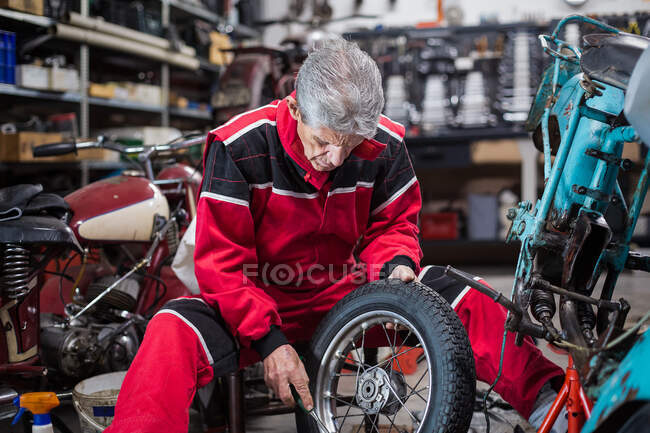Вид збоку серйозного кваліфікованого механіка літнього чоловіка в робочому одязі ремонту старого мотоцикла і кріплення колеса в майстерні — стокове фото