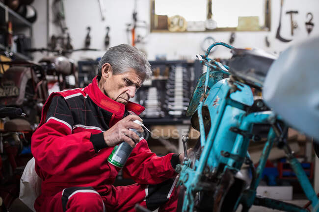 Side view of serious senior male master in workwear lubricating details of old retro motorbike with oil spray during repair work in garage — Stock Photo