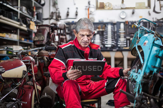 Grave cinza cabelos idosos reparador usando tablet e assistir vídeo instrução ao corrigir velho quebrado motocicleta na garagem — Fotografia de Stock