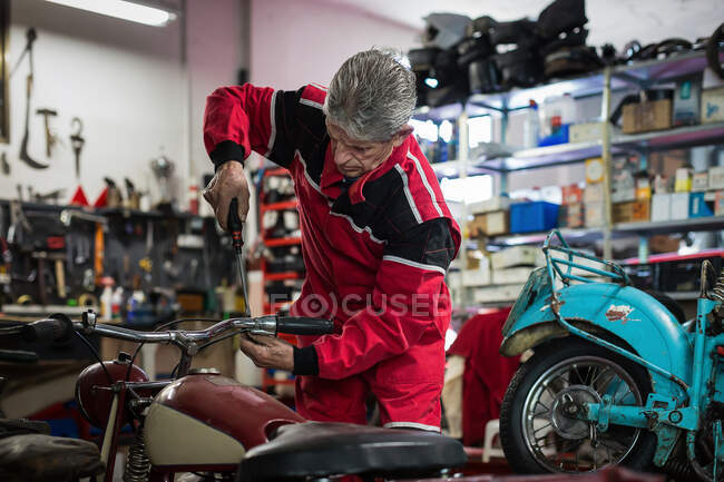 Mestre masculino sênior em vestuário de trabalho usando chave de fenda durante a fixação guidão de moto retro quebrado na oficina profissional — Fotografia de Stock