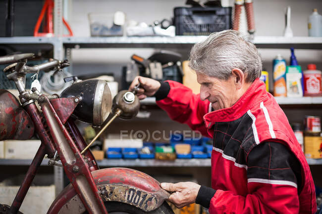Vue latérale du mécanicien masculin senior réparant une vieille moto démontée rouillée tout en travaillant dans un atelier professionnel — Photo de stock