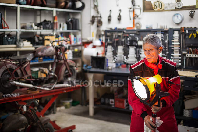 Опытный старший механик-мужчина кладет блестящую электрическую лампу рядом с рабочим местом при подготовке к ремонту в гараже с мотоциклом и инструментами — стоковое фото