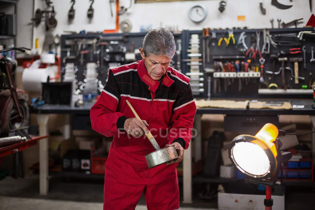 Réparateur senior qualifié en vêtements de travail mélangeant de la peinture dans un pot en métal pendant les travaux de réparation dans un atelier de service moto — Photo de stock