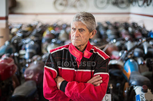 Серйозний старший чоловічий механік у червоному робочому одязі, що стоїть у майстерні ремонту проти пошкоджених іржавих мотоциклів — стокове фото