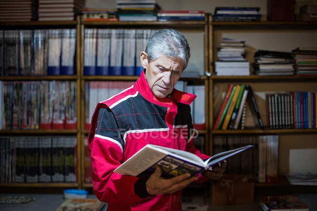 Mecánico masculino senior enfocado en libro profesional de lectura de ropa de trabajo mientras busca solución de problema técnico cerca de librería - foto de stock