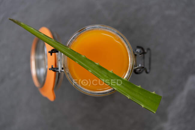 Vista superior da folha verde de aloe vera colocada no frasco de vidro cheio de suco de laranja fresco no fundo cinza — Fotografia de Stock