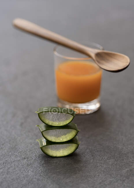 Corte pedaços de aloe vera colocados sobre mesa cinza com copo de suco de laranja e colher de madeira — Fotografia de Stock