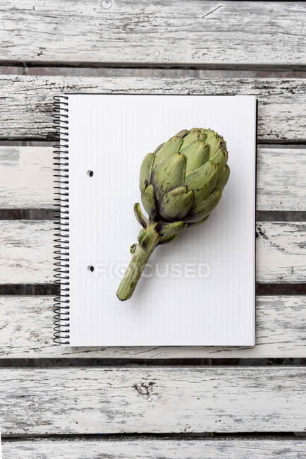Top view of green artichoke placed on opened notepad with blank pages on wooden table — Stock Photo