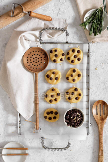 Top view of freshly baked sweet cookies with chocolate chips on metal grid placed on table with various kitchen tools and green rosemary branches — Stock Photo