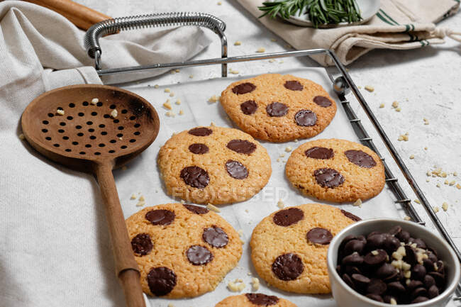 From above of freshly baked sweet cookies with chocolate chips on metal grid placed on table with various kitchen tools and green rosemary branches — Stock Photo