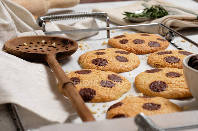 From above of freshly baked sweet cookies with chocolate chips on metal grid placed on table with various kitchen tools and green rosemary branches — Stock Photo