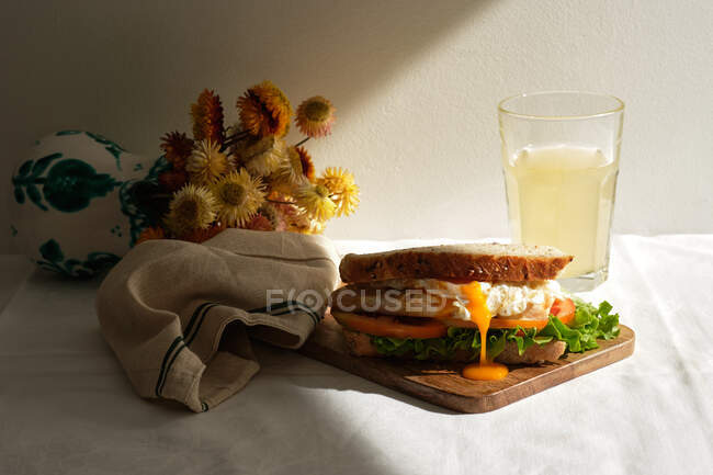 Apetitivo sándwich casero con huevos escalfados y tomates frescos y lechuga servida con un vaso de jugo para el desayuno en la mesa con flores - foto de stock