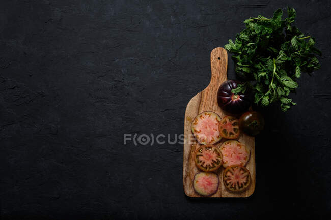 Вид сверху на свежие спелые черные помидоры и стебли зеленой мяты на деревянной разделочной доске на черном фоне — стоковое фото