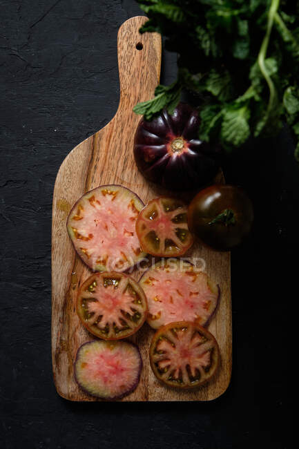 Top view of fresh ripe sliced black tomatoes and green mint stems on wooden cutting board on black background — Stock Photo