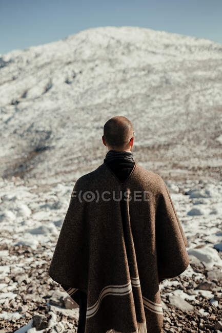Back view of anonymous male tourist in cape admiring snowy mount under blue cloudy sky in winter — Stock Photo