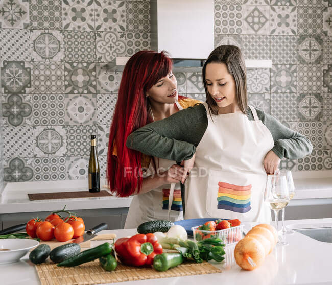 Young woman putting apron on homosexual female beloved at table with assorted fresh vegetables in house — Stock Photo