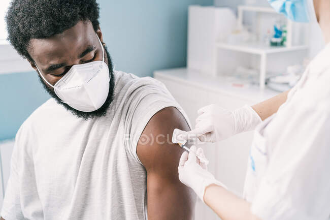 Cropped especialista médico fêmea irreconhecível em uniforme de proteção, luvas de látex e máscara facial vacinando paciente homem afro-americano na clínica durante o surto de coronavírus — Fotografia de Stock