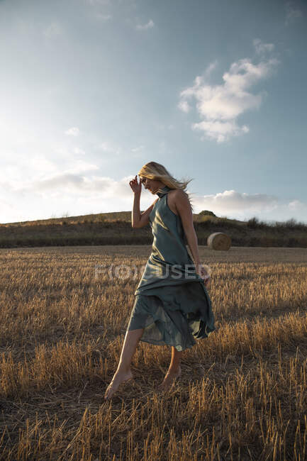 Mujer pacífica en vestido elegante de pie en el campo seco en el área rural y mirando hacia abajo - foto de stock