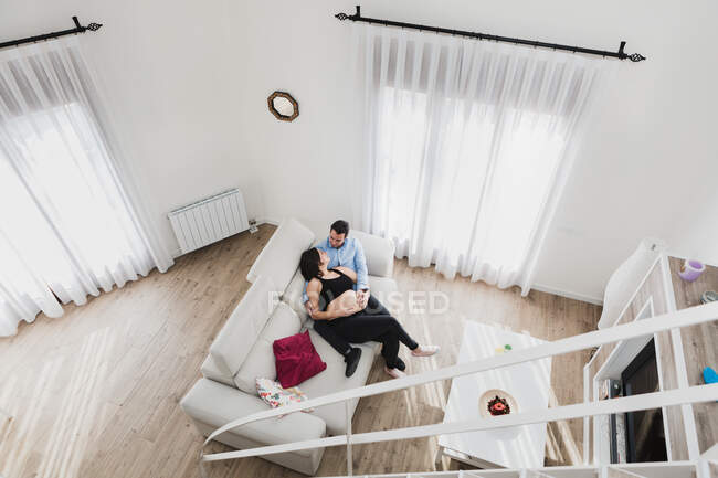 Зверху вагітна жінка сидить на дивані з чоловіком, дивлячись один на одного насолоджуючись вихідними разом вдома — стокове фото