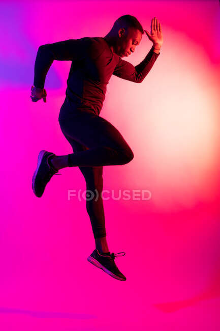 Visão lateral de corpo inteiro do atleta masculino afro-americano em sportswear pulando na iluminação de néon roxo e rosa do estúdio — Fotografia de Stock