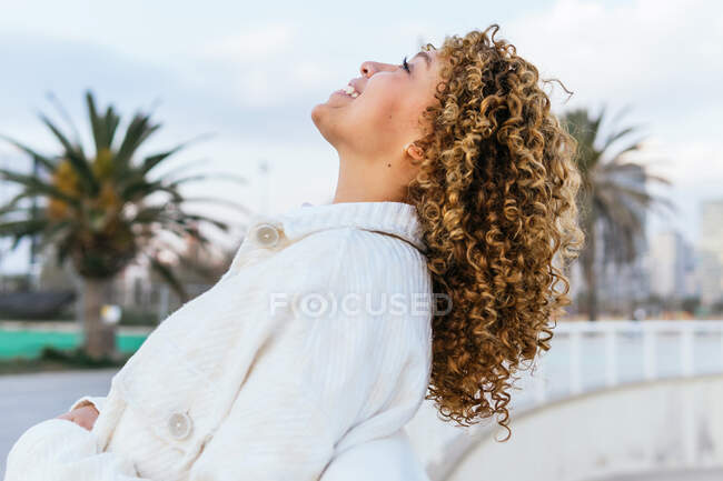 Seitenansicht einer unbeschwerten Afroamerikanerin mit lockigem Haar, die am Zaun lehnt und abends in der Stadt aufblickt — Stockfoto