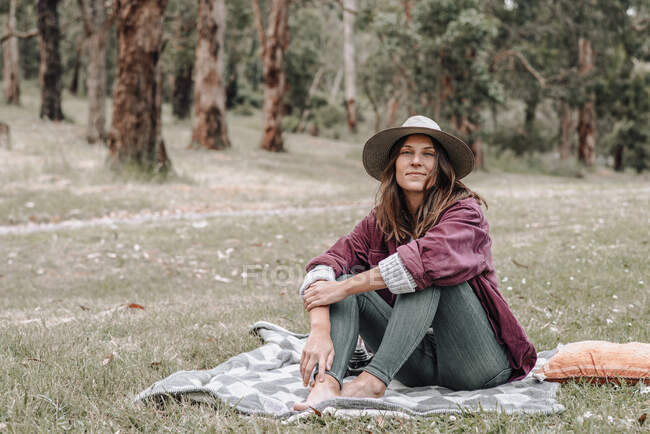 Cheerful female sitting on plaid on meadow in woods and looking at camera while enjoying picnic in Australia — Stock Photo