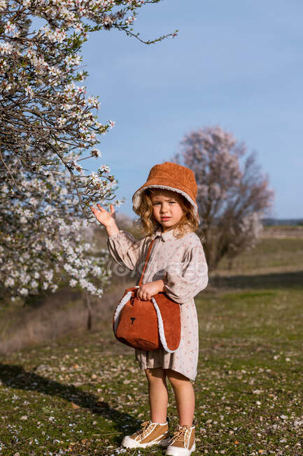Entzückendes kleines Mädchen in Kleid und Hut steht am Baum mit blühenden Blumen und blickt in die Kamera im Frühlingsgarten — Stockfoto