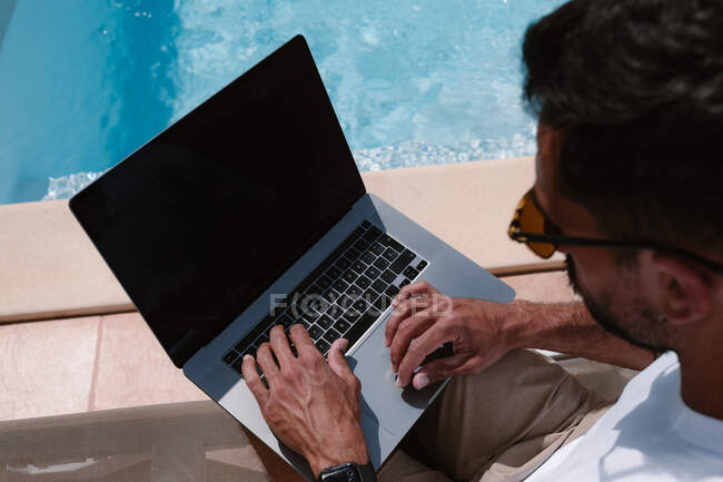 Male freelancer lying on lounger at poolside and surfing Internet on laptop during telework in summer on sunny day — Stock Photo