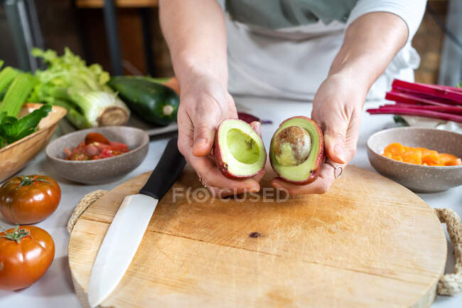 Cultivo femenino anónimo con mitades de aguacate maduras por encima de la tabla de cortar durante el proceso de cocción en casa - foto de stock