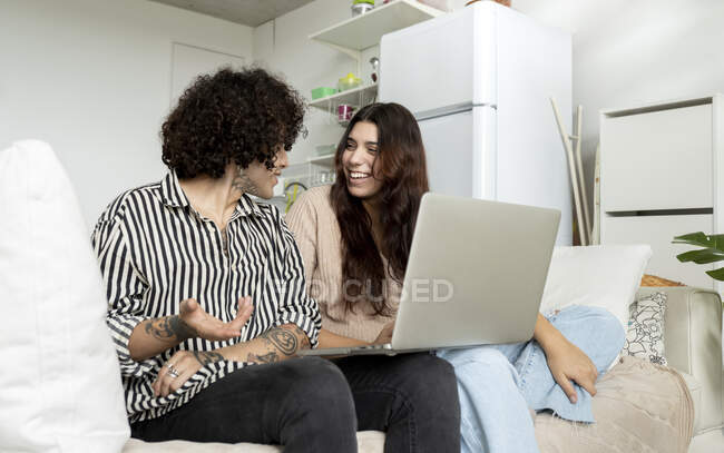 Fröhliche Frau mit Netbook spricht mit tätowiertem Freund, während sie sich auf der Couch im Hauszimmer anschaut — Stockfoto