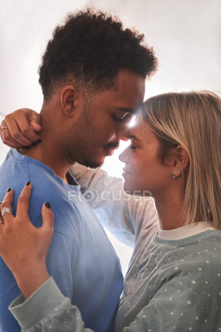 Seitenansicht des ruhigen multirassischen Paares im Pyjama, das sich morgens zu Hause von Angesicht zu Angesicht umarmt — Stockfoto