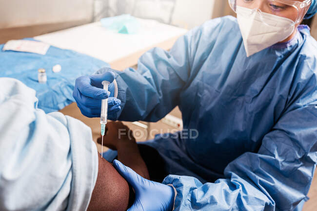 Cultivé médecin féminin en uniforme de protection et gants en latex vaccinant patient afro-américain masculin méconnaissable en clinique pendant l'épidémie de coronavirus — Photo de stock